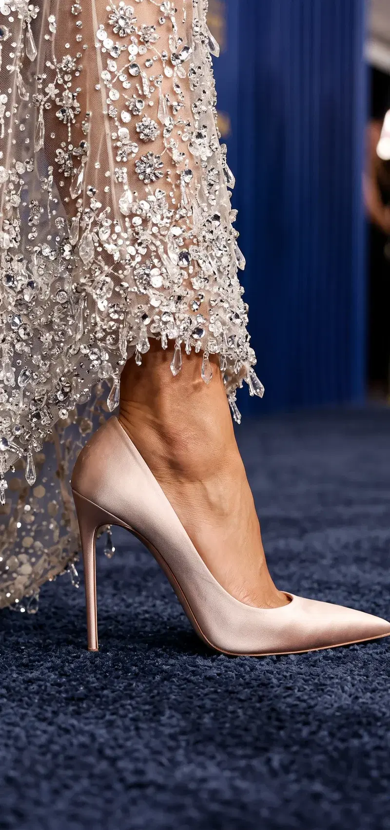 Pale rose satin pointed-toe stiletto pump in profile under a crystal-fringe gown hem on a navy carpet